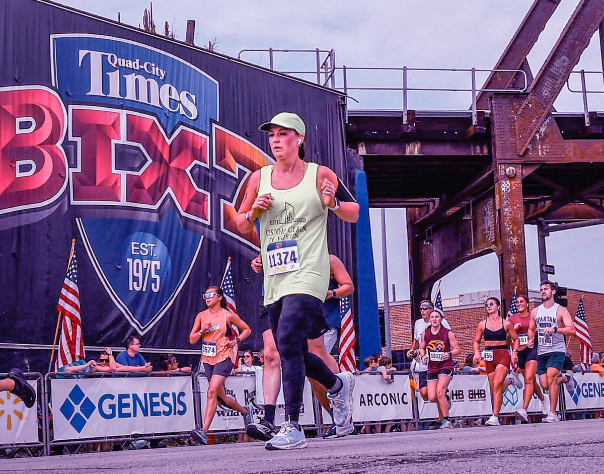 Photo of Rachel Loussaert running on asphalt in the Bix 7 marathon in Davenport, Iowa, with part of a rusted bridge, and other runners in the background.